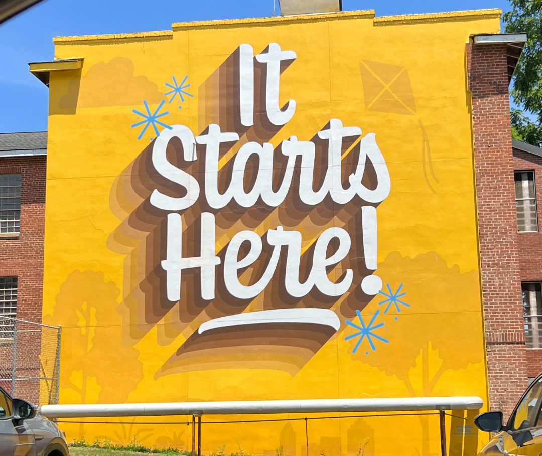 yellow sign on a building in Raleigh with bold cursive font It starts here